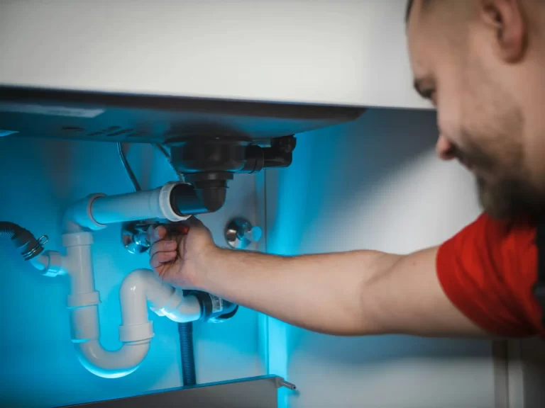 A plumber in a red shirt working on pipes under a sink